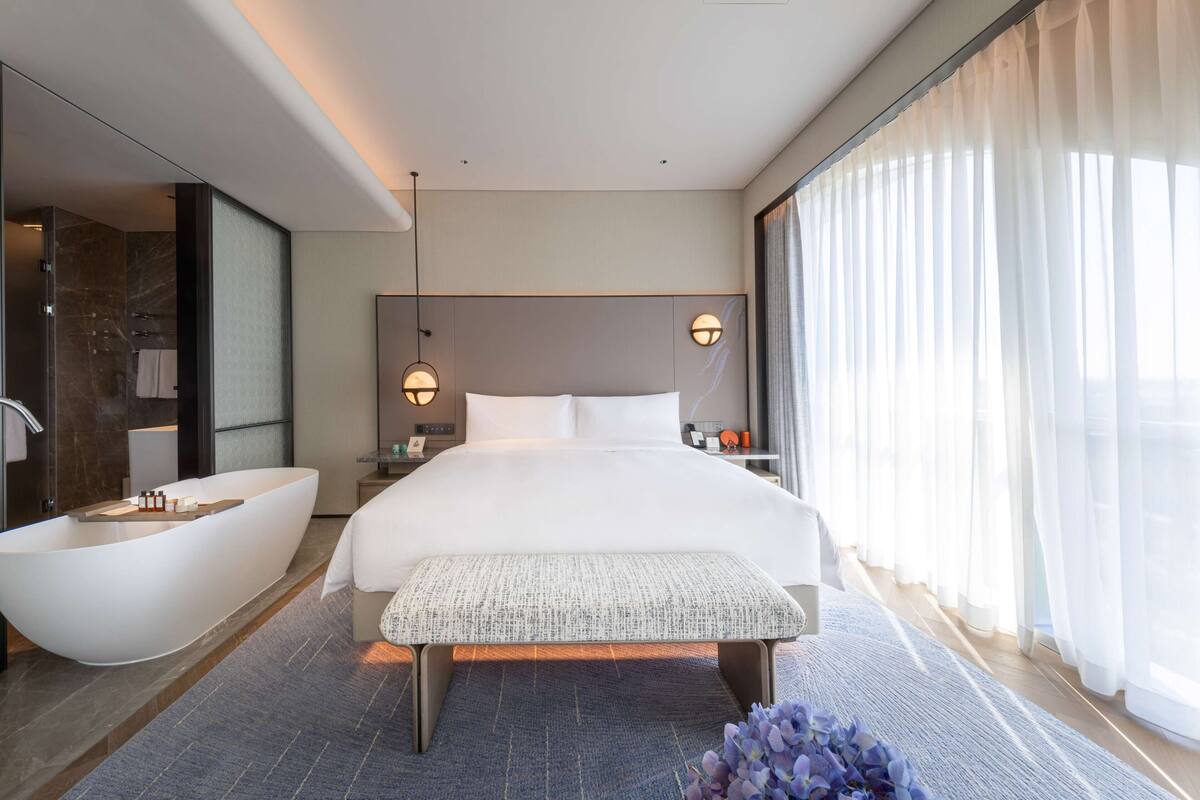 A modern hotel room with a large bed, a built-in bathtub, a bench, and a large window with sheer curtains.