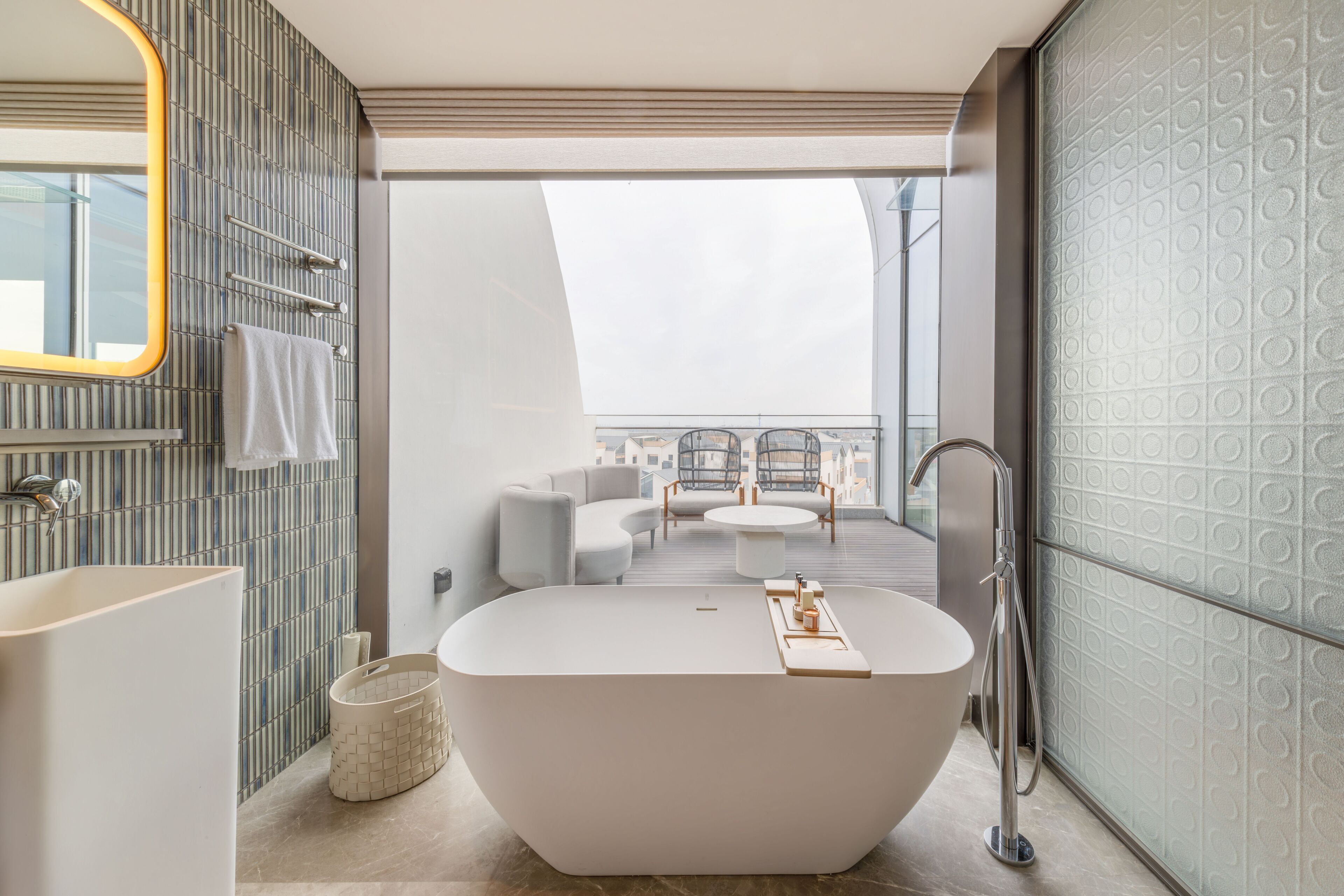 A modern bathroom with a freestanding bathtub, a large mirror, and a view of a balcony with outdoor furniture.