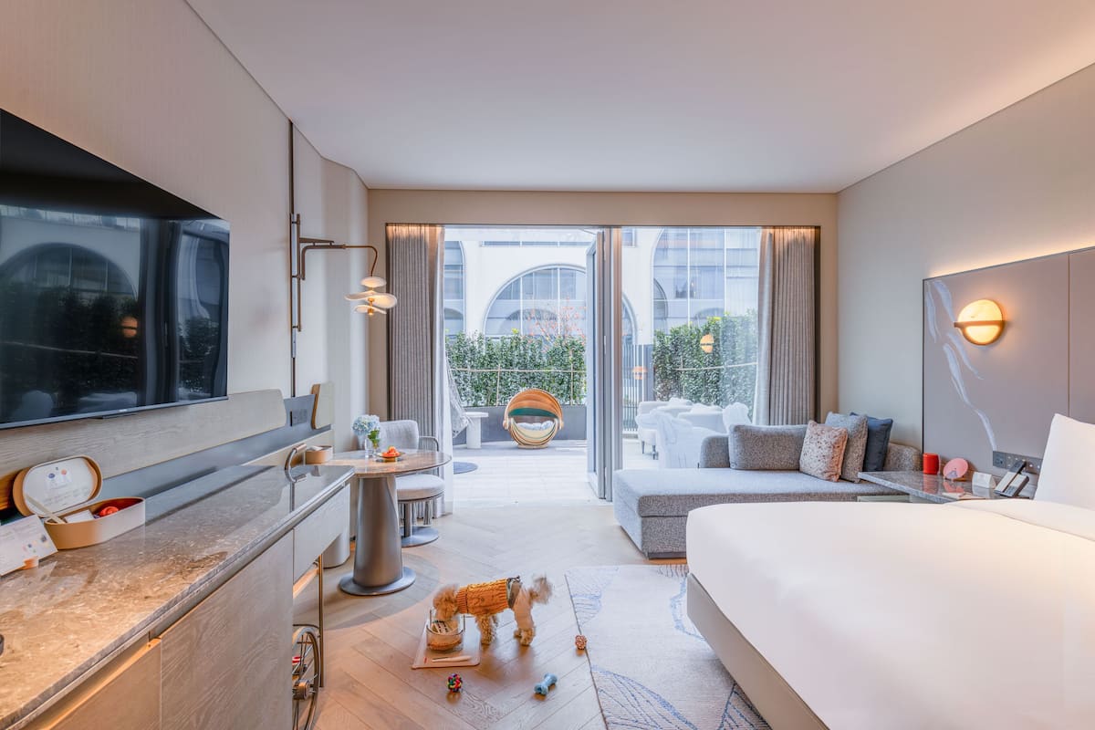 A modern hotel room with a large bed, a desk with a computer, a small table, a sofa, and a balcony with outdoor seating.
