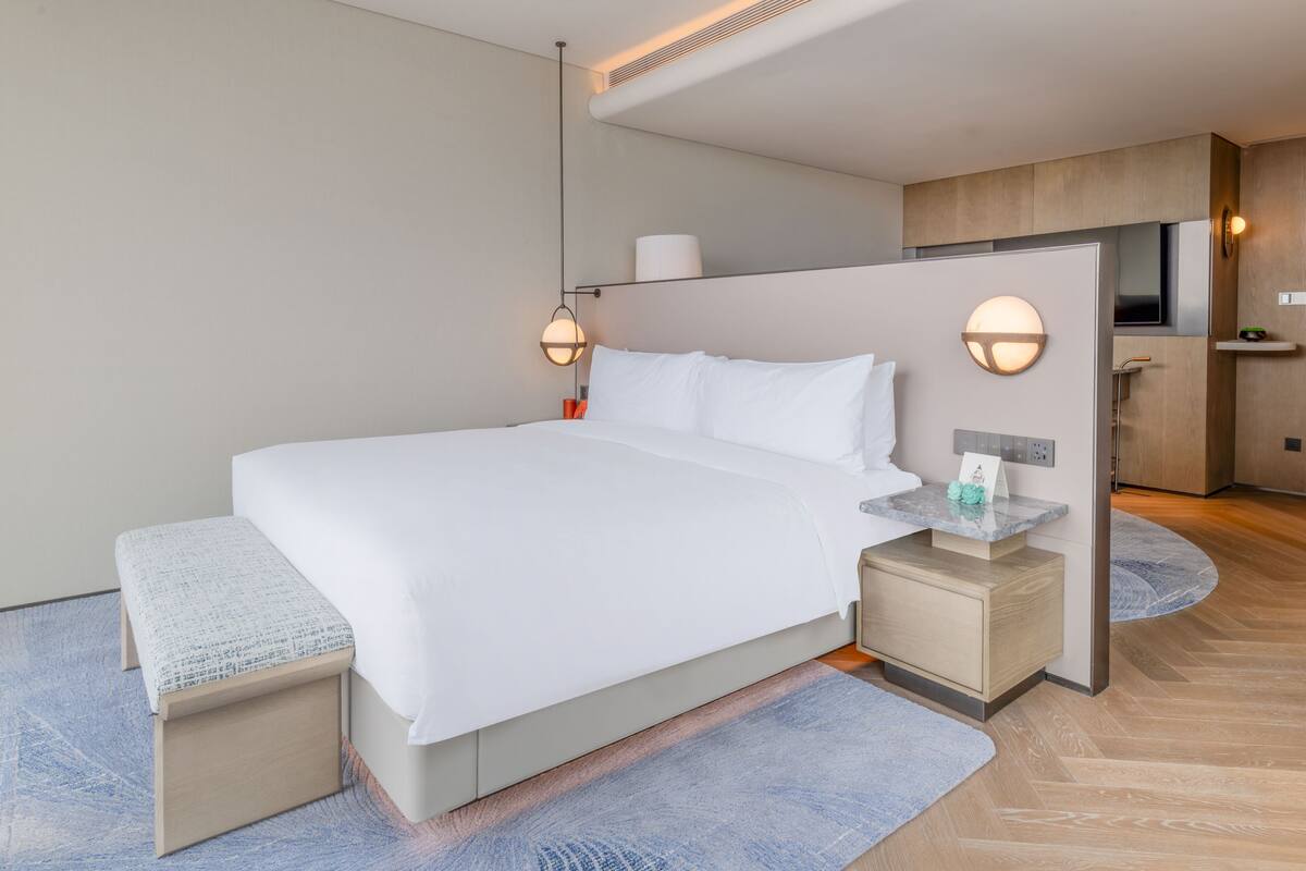 A modern hotel room with a large bed, bedside tables, a small desk, and a built-in TV.