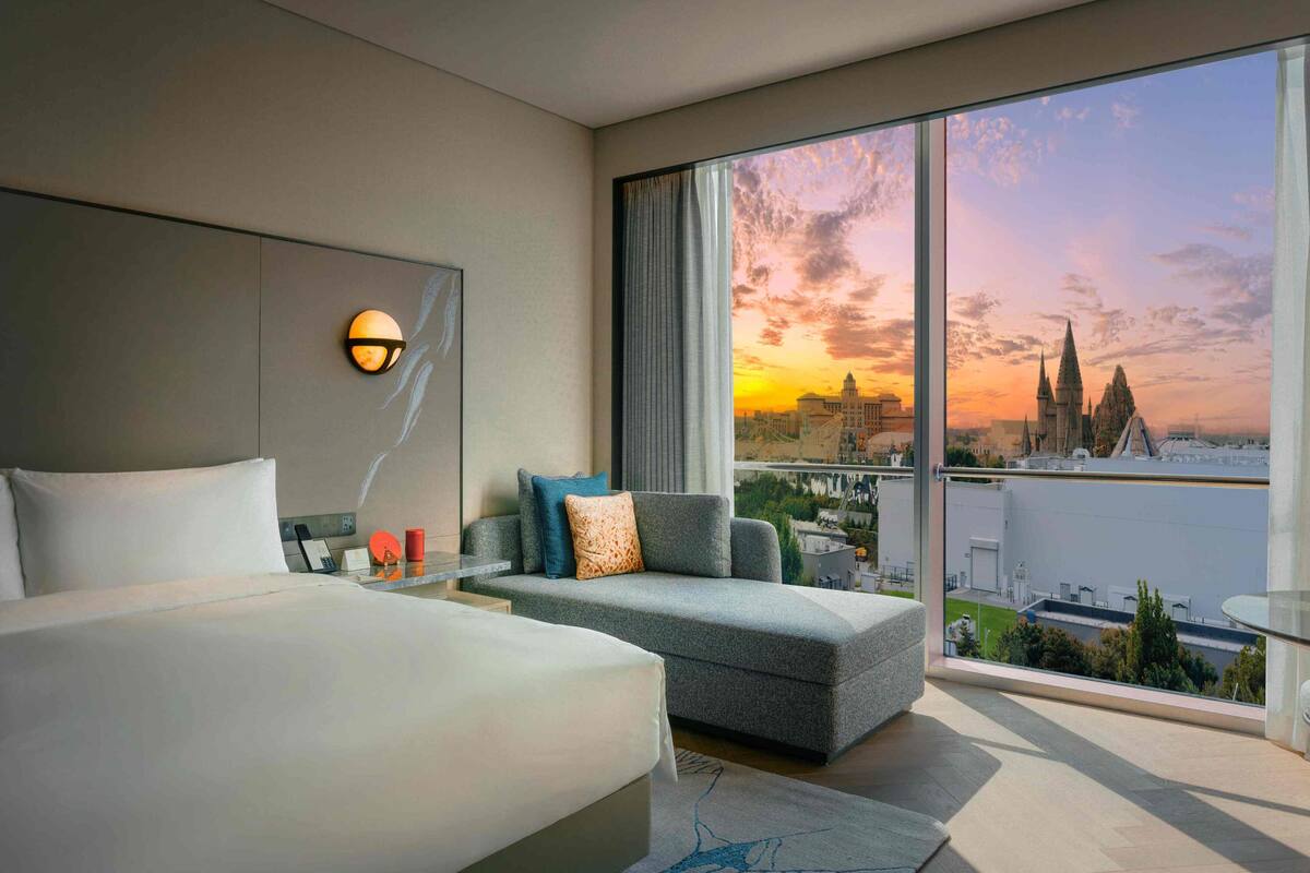 A modern hotel room with a large bed, a sofa, and a view of a cityscape at sunset.
