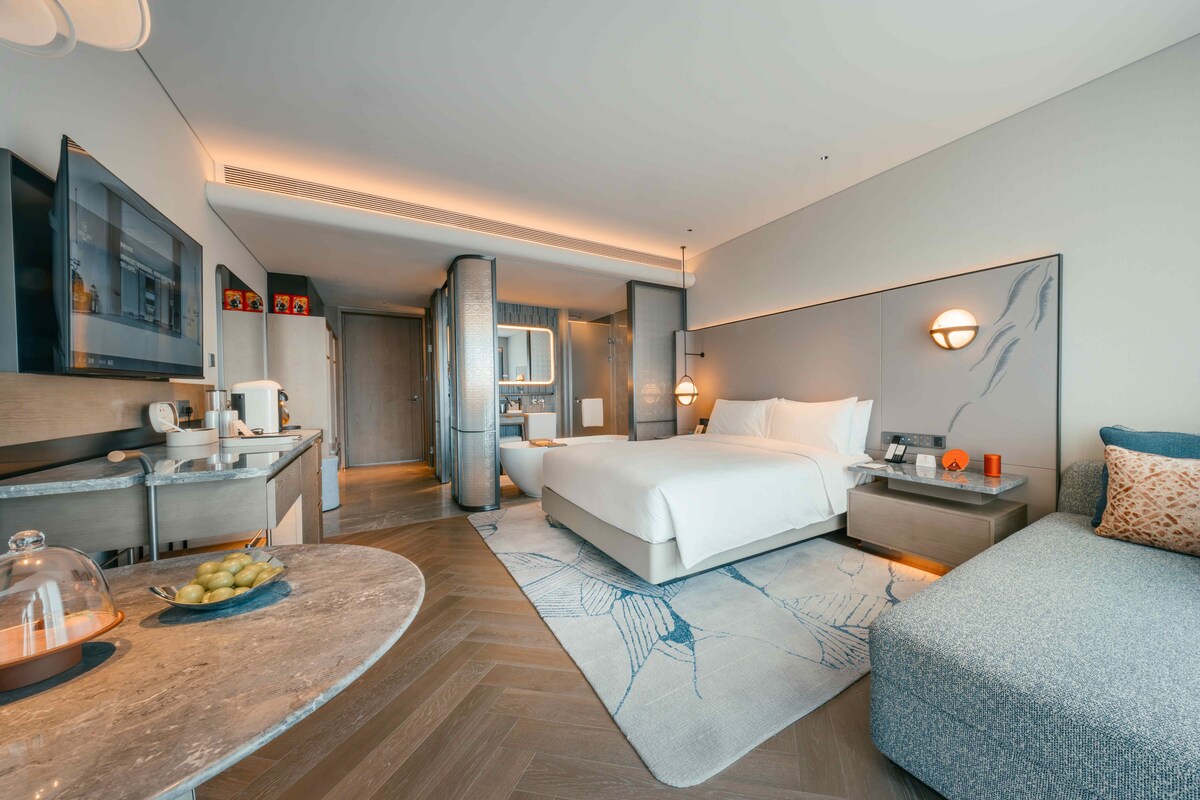Habitación de hotel moderna con una cama grande, un escritorio con computadora, una zona de estar y un baño visible a través de una puerta abierta.
