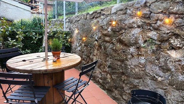 An outdoor patio with a round wooden table and black chairs, a black grill, and string lights against a stone wall. - La Voltina – The ideal starting point for exploring Lucca and the Garfagnana (Diecimo)