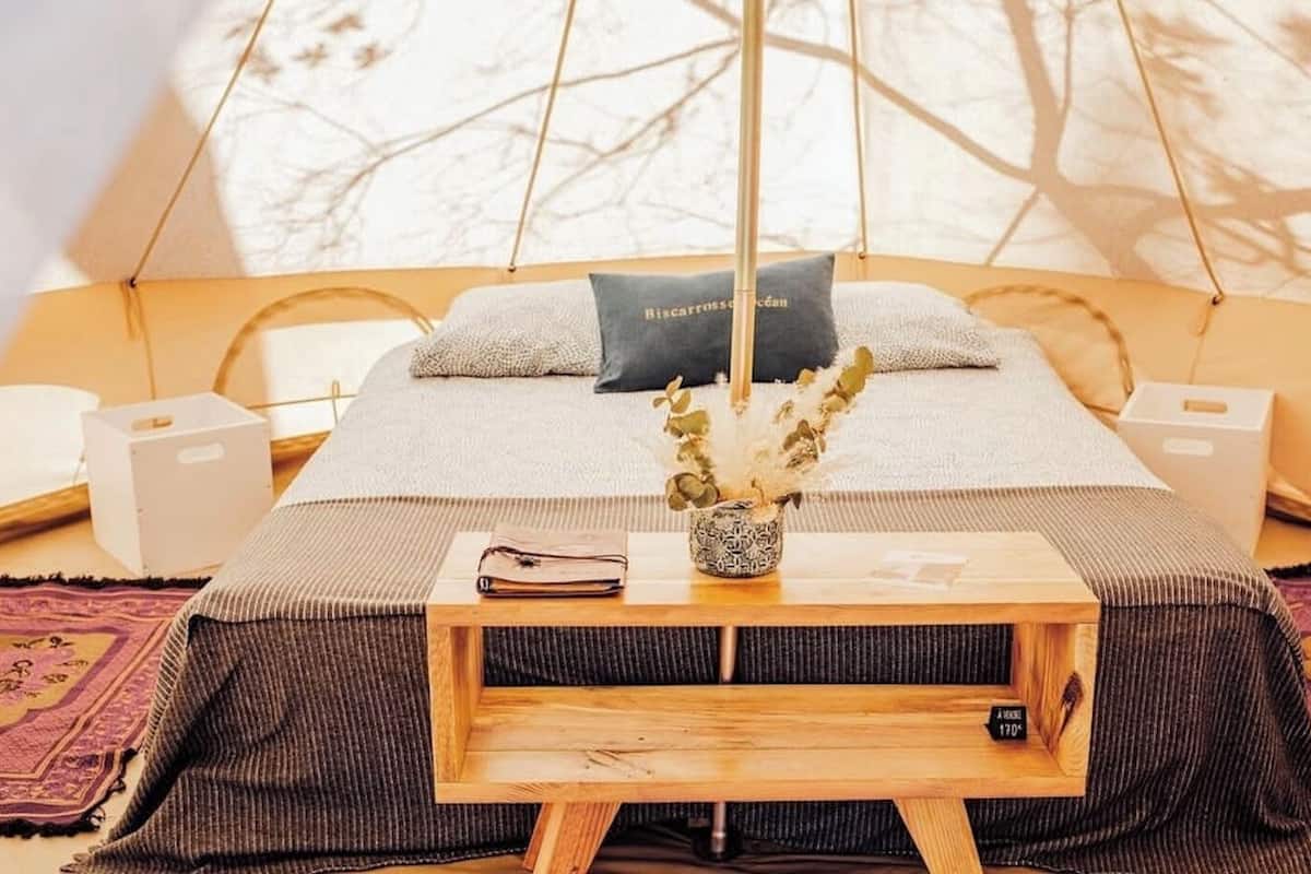 A cozy room with a bed, a bedside table with a book and a vase, and a view of trees through a window.