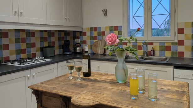 A kitchen with white cabinets, a colorful tiled backsplash, a wooden table with glasses and a bottle, and a window with a view. - Window on castle (Monflanquin)