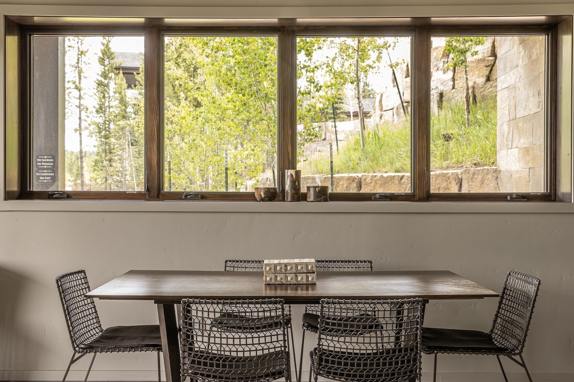 A dining area with a table and wire mesh chairs, a window with a view of trees and a stone wall, and a "No Smoking" sign on the window frame.