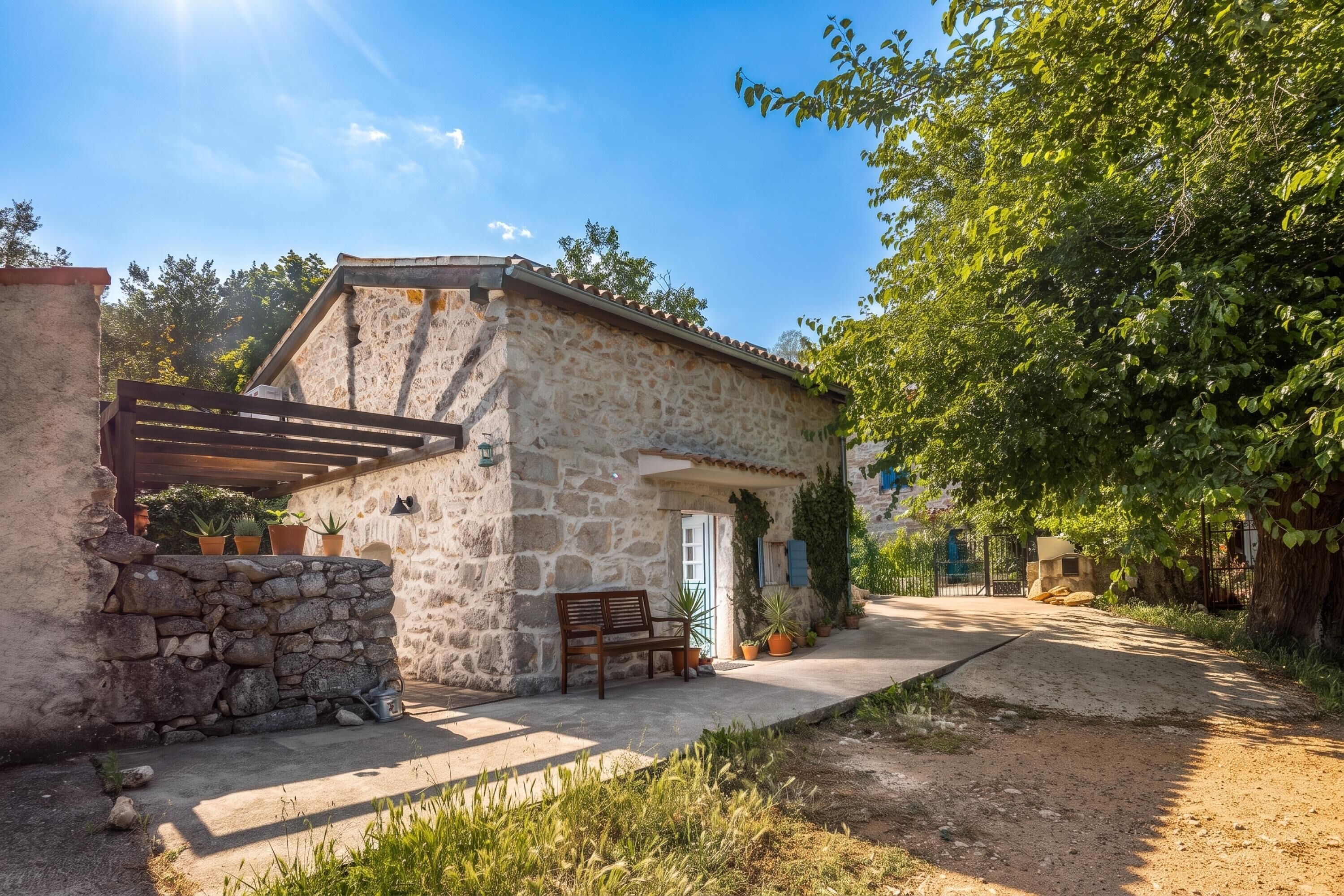 A stone building with a wooden bench outside, a stone wall, and a paved path leading to a gate.