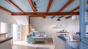 A cozy living space with a high ceiling, wooden beams, a grey sofa, a small table with a lamp, and a staircase leading to an upper level.