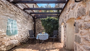 A stone-walled courtyard with a wooden pergola, a table with chairs, and a hanging cloth.