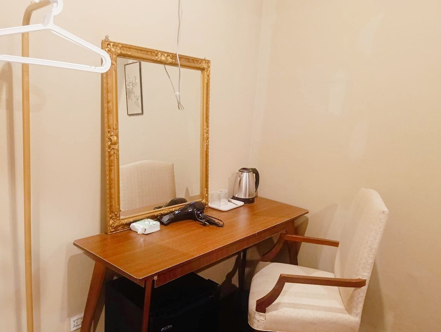 A room with a wooden desk, a chair, a mirror with a gold frame, a black hairdryer, and a kettle on the desk.