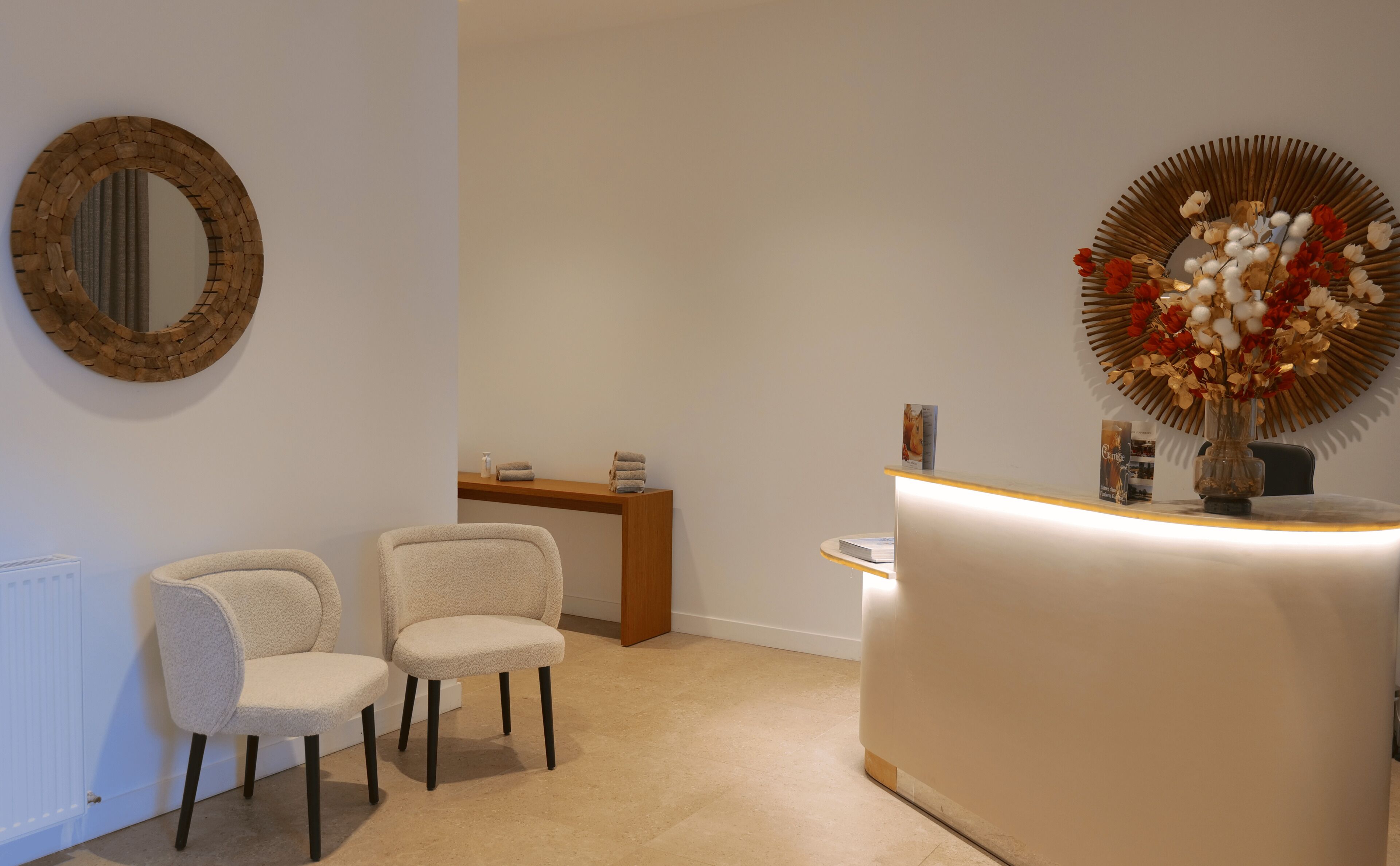 Una zona de recepción con un escritorio de madera, dos sillas, un espejo y un jarrón decorativo con flores.