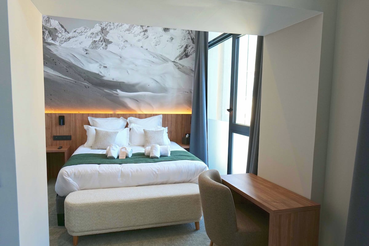 A modern hotel room with a large bed, a wooden desk, and a mountain mural on the wall.