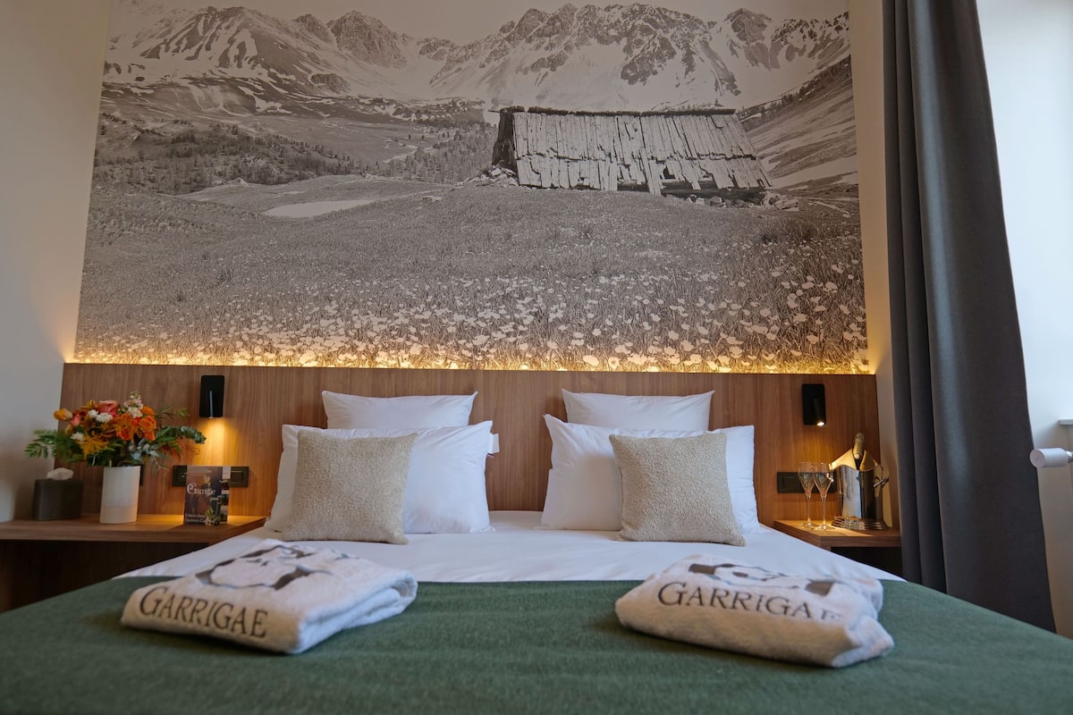 A hotel room with a bed, pillows, a nightstand with a lamp, a wall-mounted artwork, and a window with curtains.