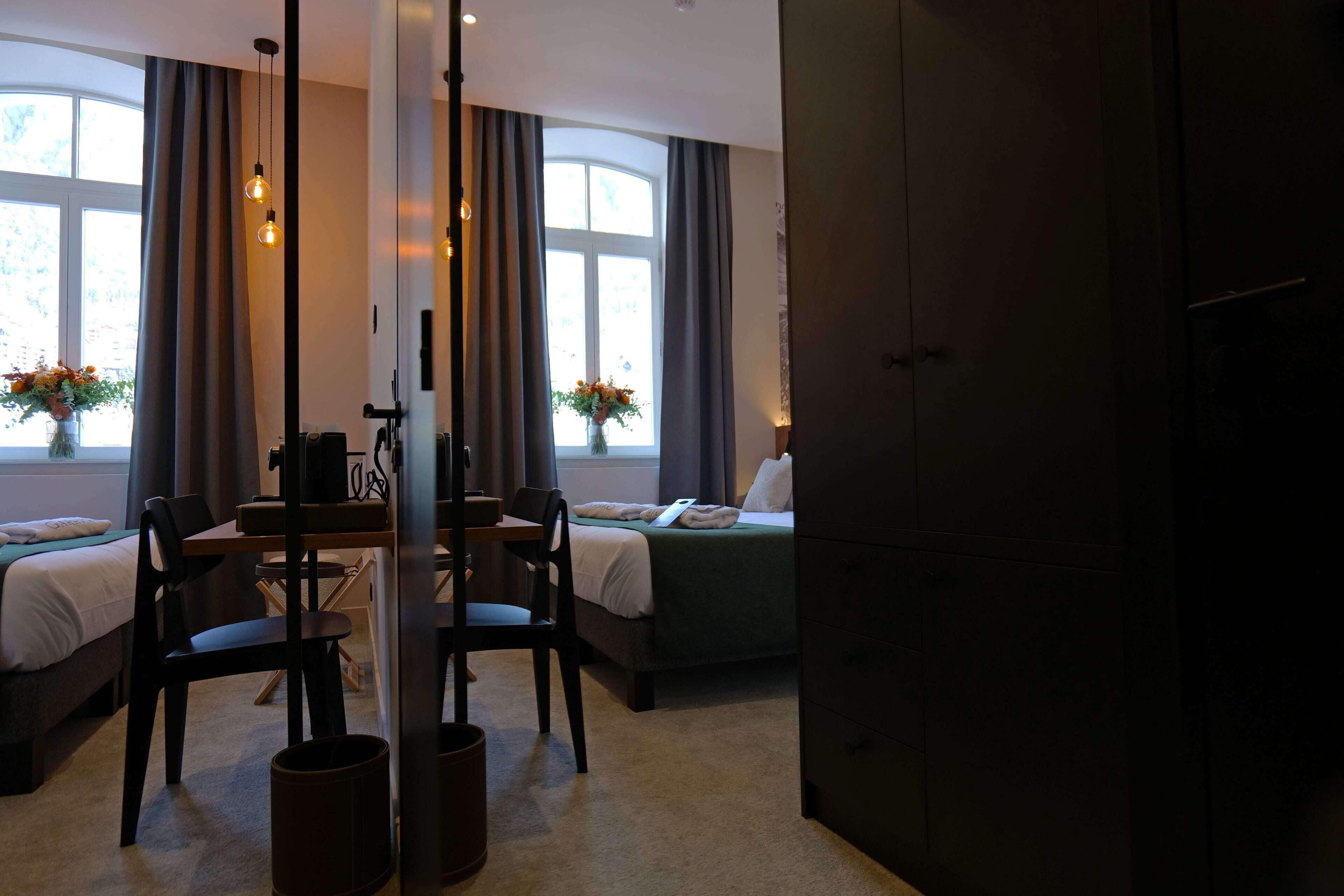 A hotel room with a bed, a desk with chairs, a vase with flowers, and a window with curtains.