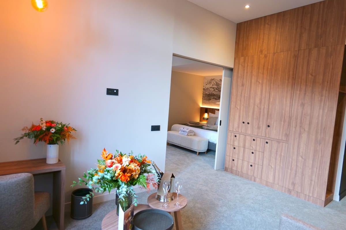 A modern hotel room with a wooden wardrobe, a small seating area with a table and chairs, and a bed with a mountain view.