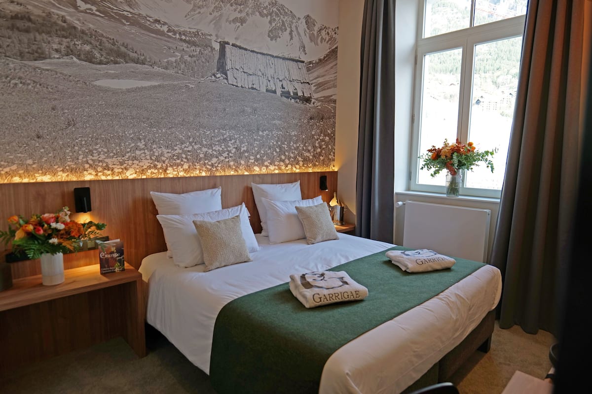 A hotel room with a large bed, a wooden headboard, a nightstand with a vase of flowers, and a window with curtains.
