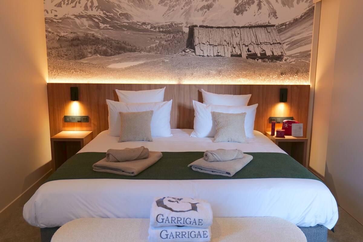 A hotel room with a large bed, two bedside tables, a wall-mounted lamp, and a mountain landscape mural.