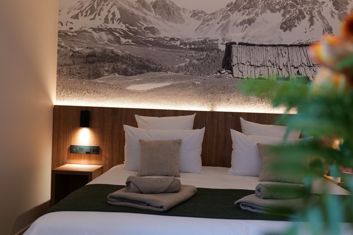 A neatly made bed with a mountain landscape mural above it, a bedside table with a lamp, and a wooden headboard.