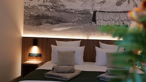 A neatly made bed with a mountain landscape mural above it, a bedside table with a lamp, and a wooden headboard. - Garrigae Caserne de Briançon & Spa - les Cinq Mondes (Briançon)