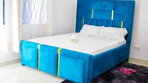 A bed with a blue headboard and white bedding, a small bedside table, and a black furry rug. - Fully furnished onebedroom in Mombasa Bamburi Vescone1 estate (Mombasa)