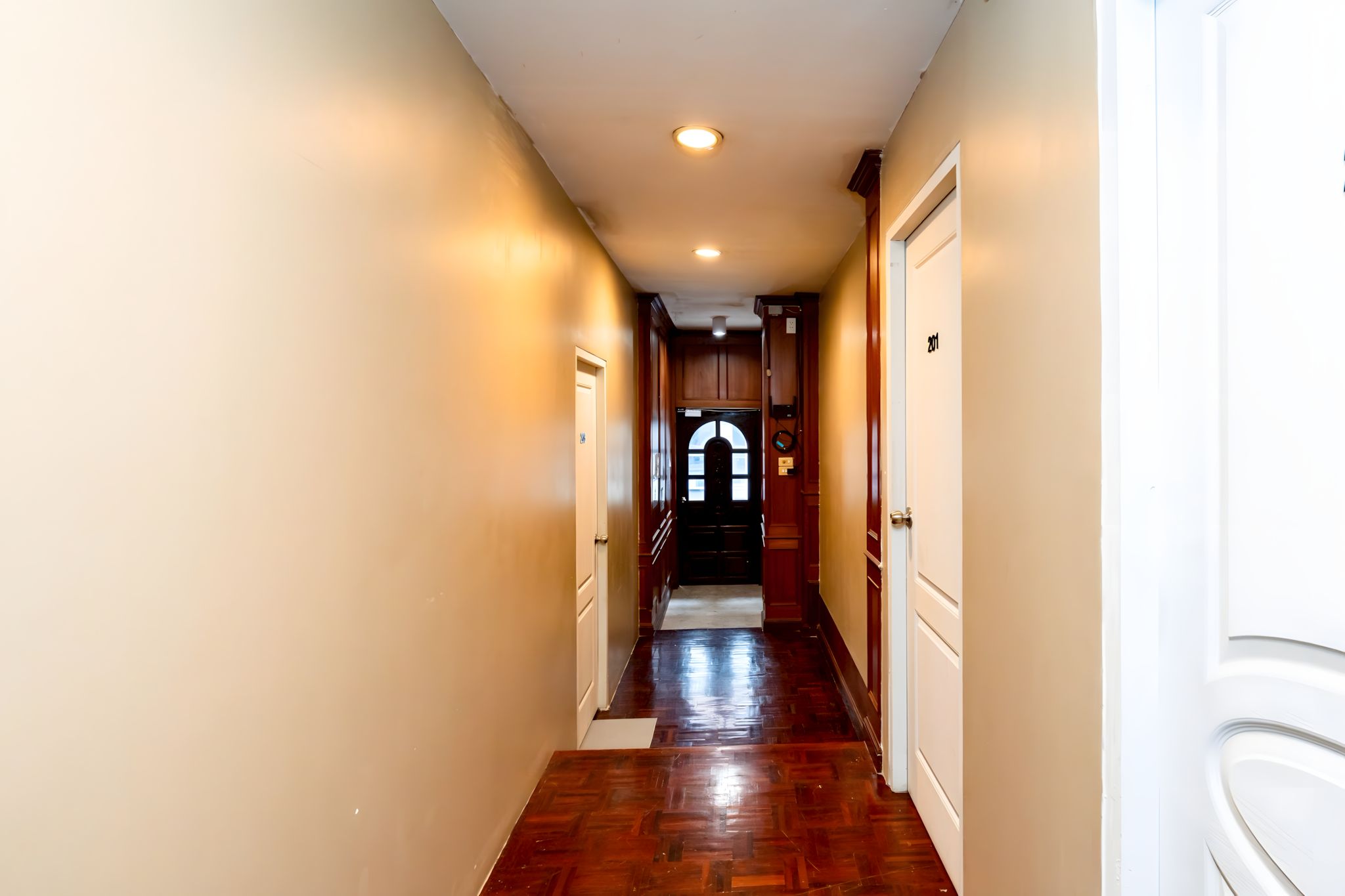 A hallway with wooden floors, white walls, and a door at the end leading to a room.