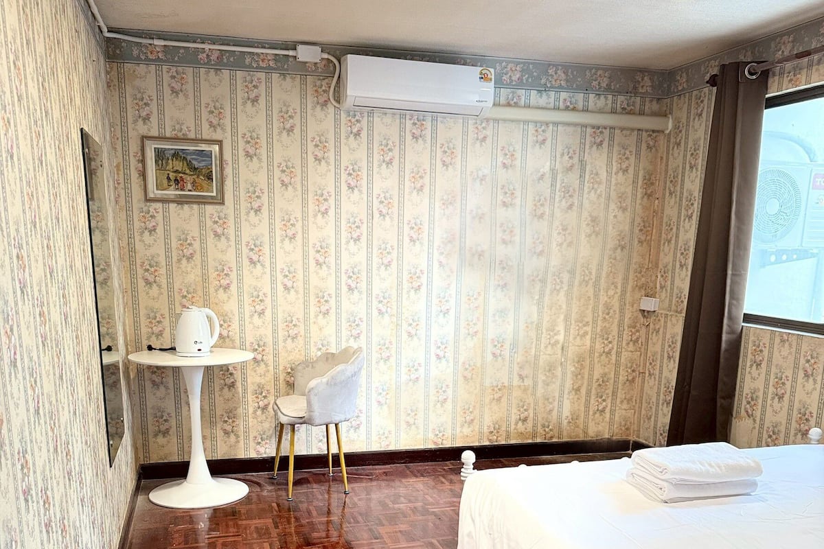 A hotel room with a wooden parquet floor, a bed with white bedding, a small round table with a chair, and floral wallpaper.