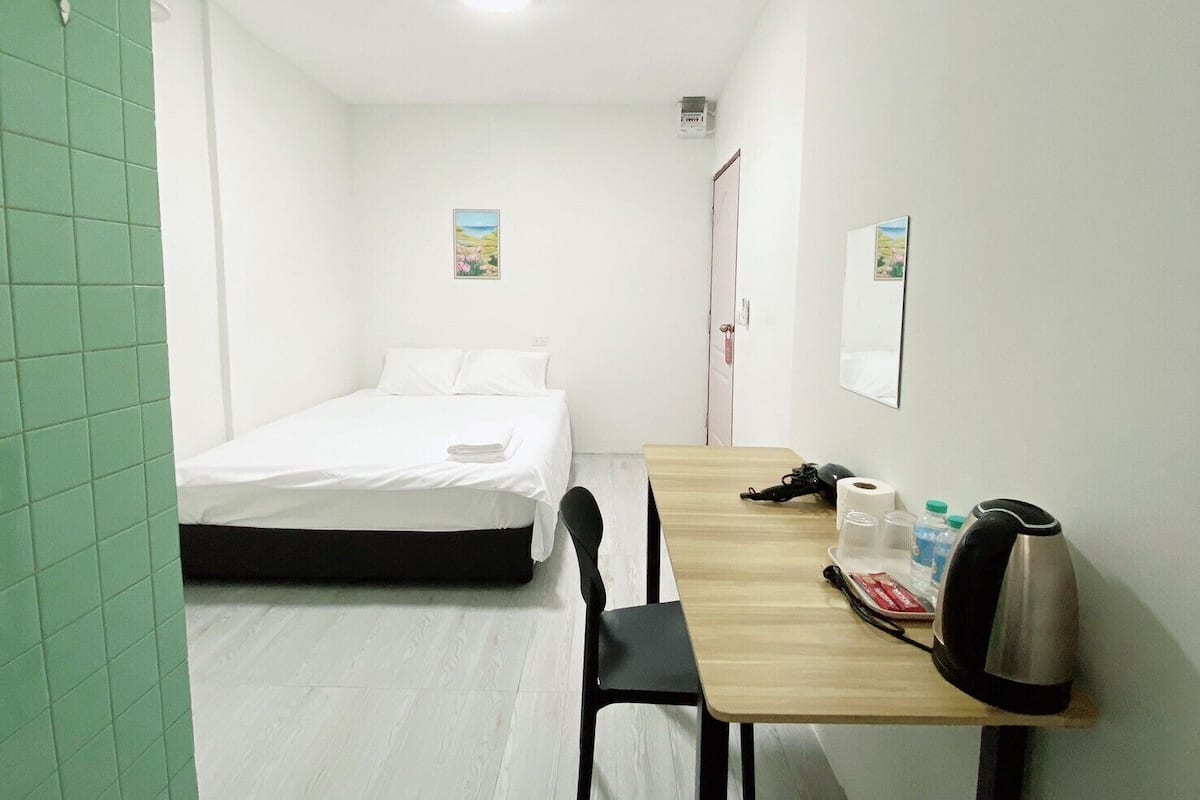 Quarto de hotel com uma cama, uma mesa de madeira com chaleira, uma cadeira e porta para outro quarto.