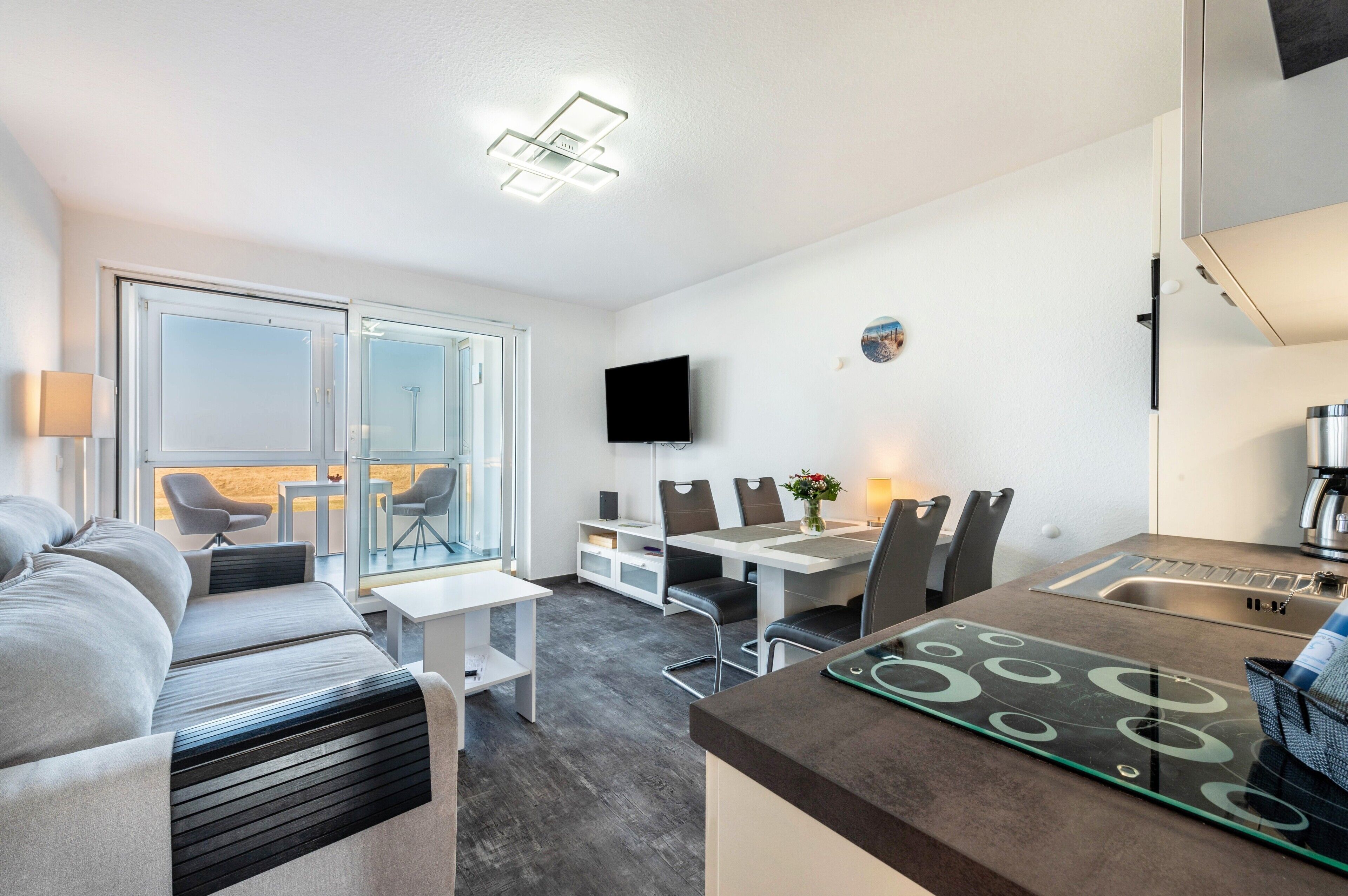 A modern hotel room with a sofa, a dining area with chairs and a table, a kitchen with a sink, and a TV mounted on the wall.