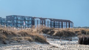 A coastal building with multiple windows, a sandy foreground with grass, and a clear sky.