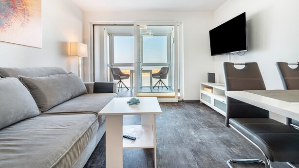 A modern hotel room with a sofa, a small table, a TV, and a balcony with chairs.