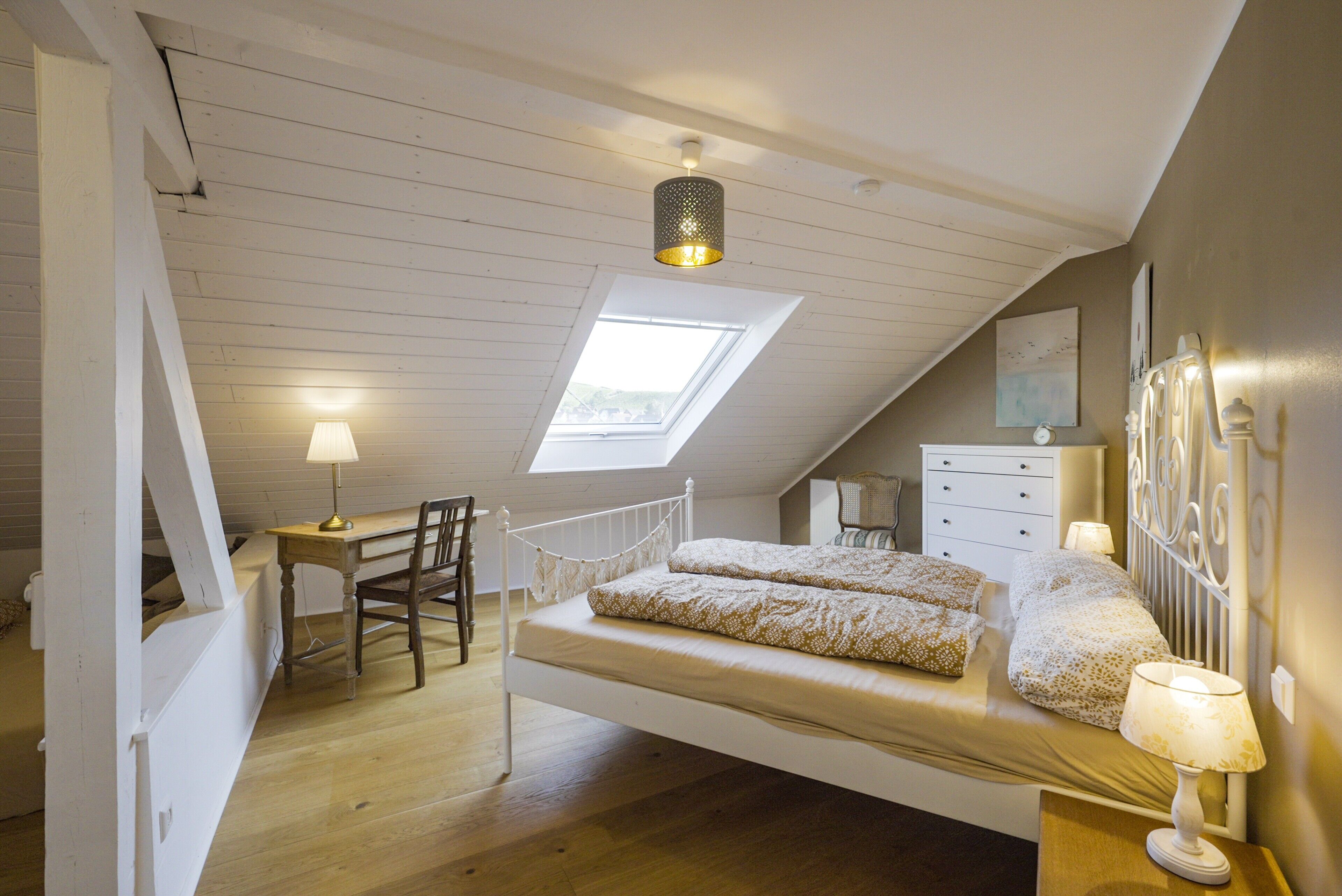 Een slaapkamer met een schuine kap, dakraam, bed met gestreept beddengoed, nachtkastje met lamp, stoel en een dressoir.