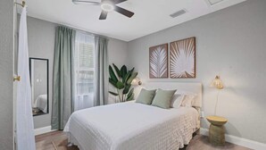 Ein Schlafzimmer mit Bett, Deckenventilator, Fenster mit Vorhängen und Wandbildern.