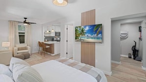 A modern hotel room with a bed, a desk, a kitchenette, and a painting of a beach scene.
