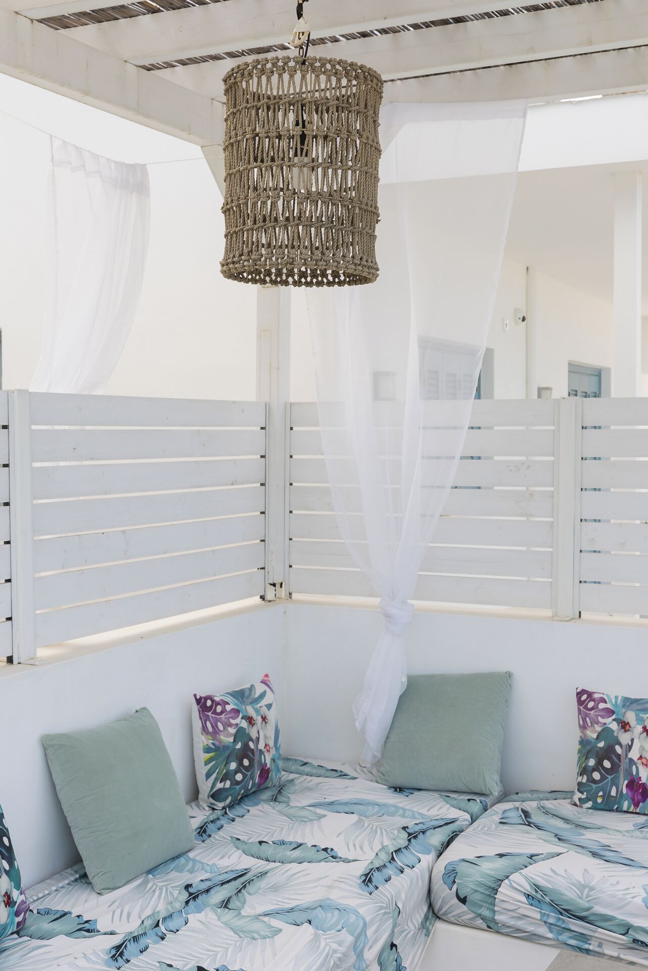 Une pièce avec un luminaire suspendu au plafond, dont l’abat-jour est fait de fibres tressées, des rideaux blancs et un lit recouvert d’une literie à motifs floraux.