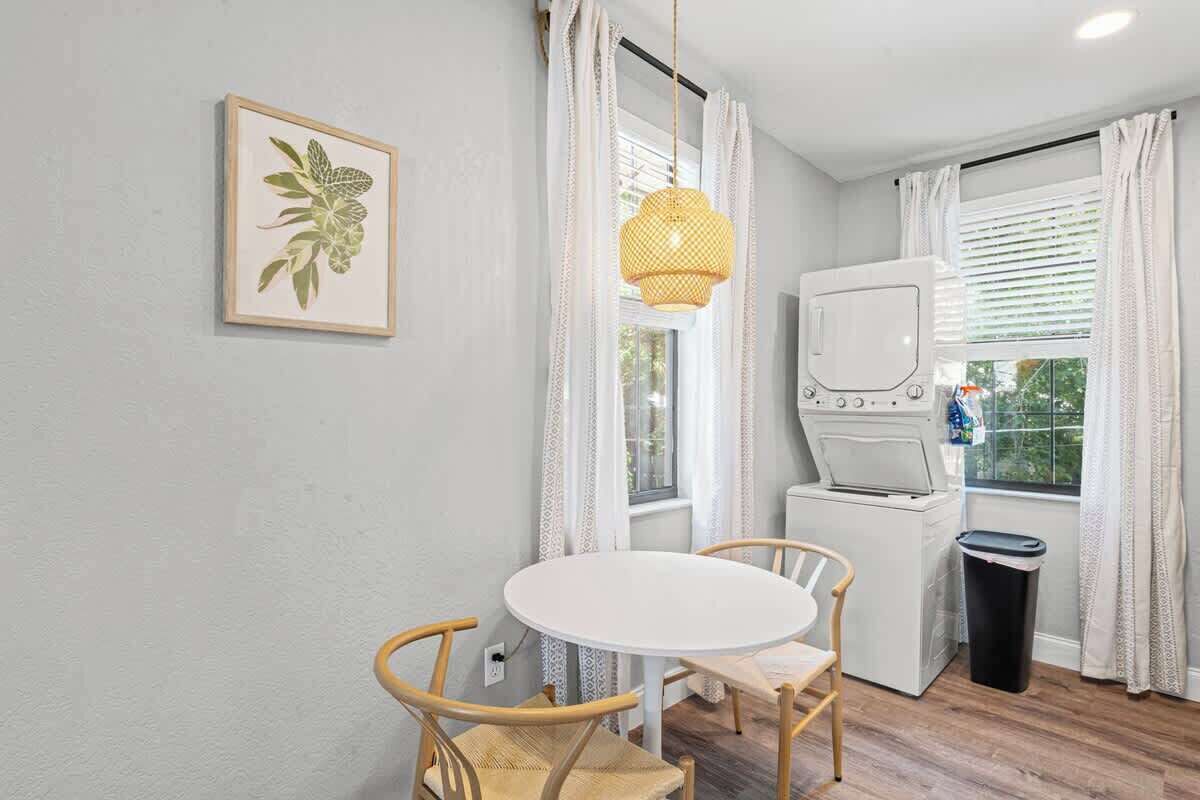 Una habitación con una mesa redonda, dos sillas, una lavadora, una lámpara colgante y un cuadro en la pared.