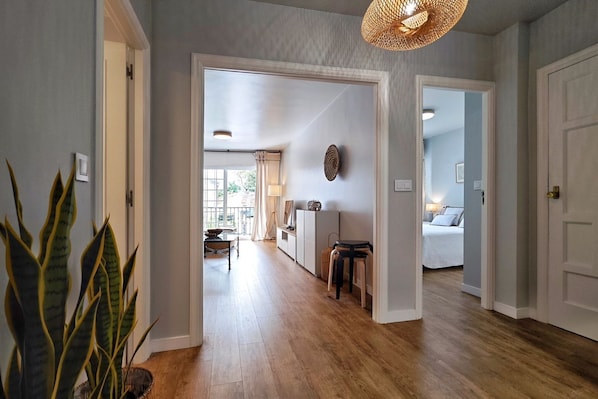 A hallway with a wooden floor, a plant, a woven light fixture, and a view into a bedroom with a bed and a window.