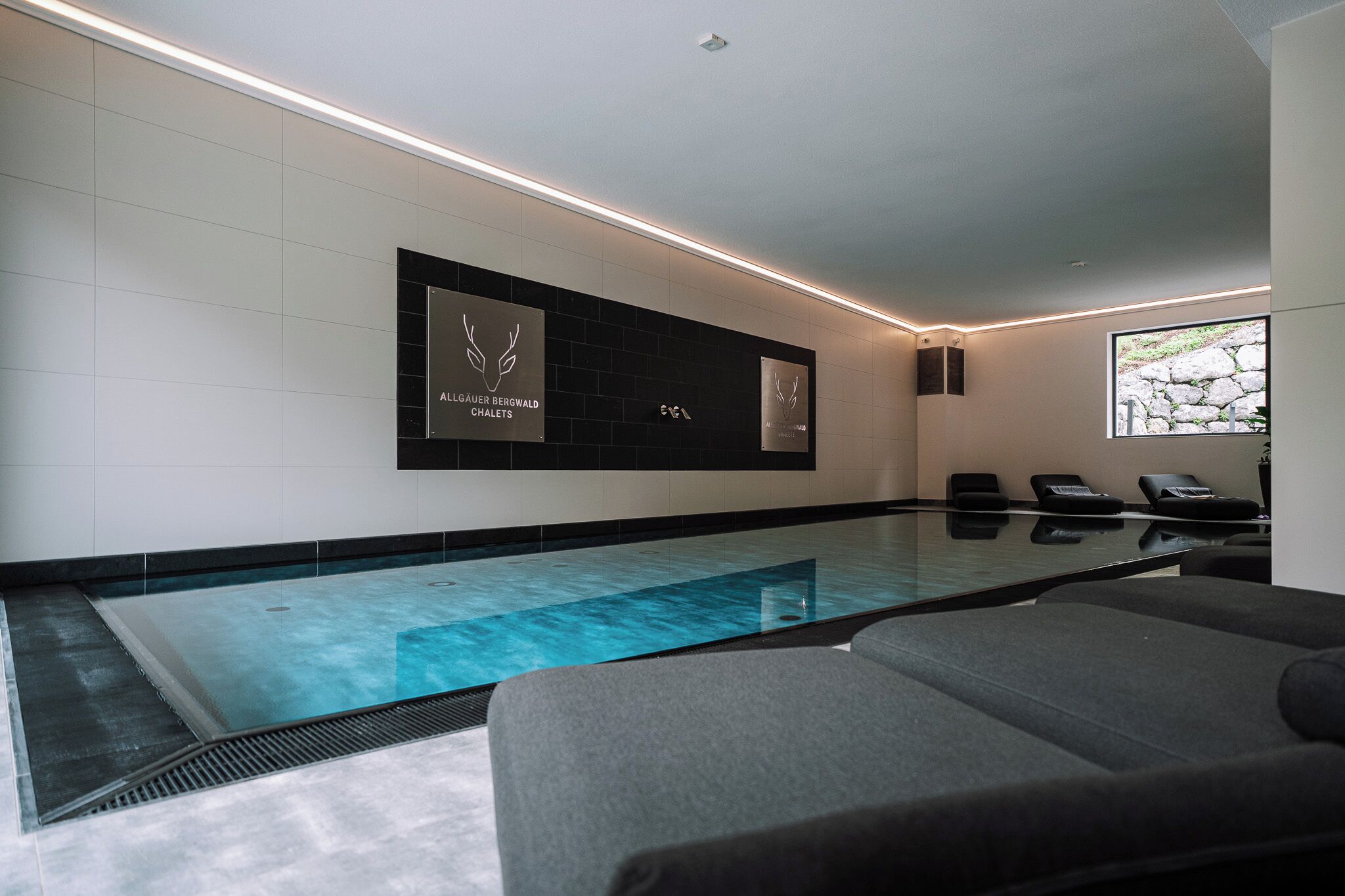 A modern indoor pool area with a clear pool, lounge chairs, and a wall-mounted sign.