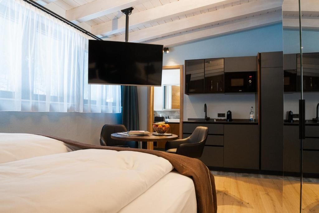 Ein Hotelzimmer mit Bett, Fernseher, einem kleinen Esstisch mit Stühlen, einer Küchenzeile und einem großen Fenster mit durchsichtigen Vorhängen.