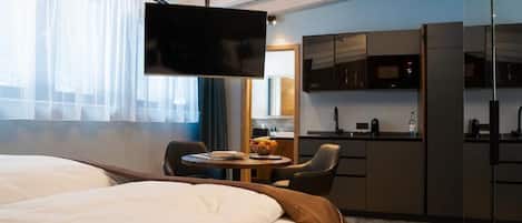 Une chambre d’hôtel comprenant un lit, une télévision, une petite table à manger avec des chaises, un coin cuisine et une grande fenêtre avec des rideaux légers.