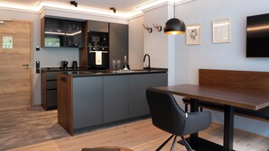 A modern kitchen with a built-in island, a dining area with a wooden table and black chairs, and a living space with a flat-screen TV and artwork on the wall. - Allgäuer Bergwald Chalets  (Oberstaufen)