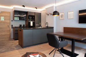 A modern kitchen with a built-in island, a dining area with a wooden table and black chairs, and a living space with a flat-screen TV and artwork on the wall. - Allgäuer Bergwald Chalets  (Oberstaufen)