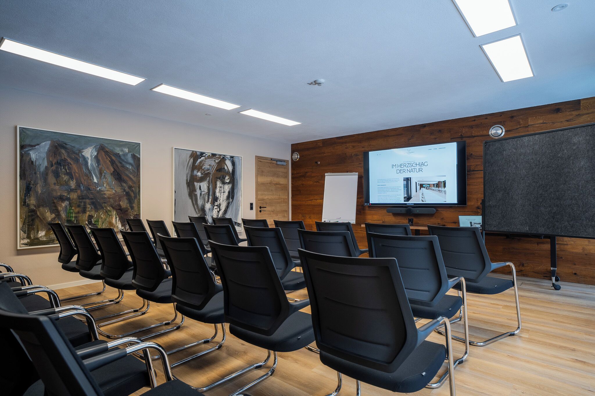 Ein Konferenzraum mit Reihen schwarzer Stühle, einer großen Leinwand und Holzvertäfelung an den Wänden.
