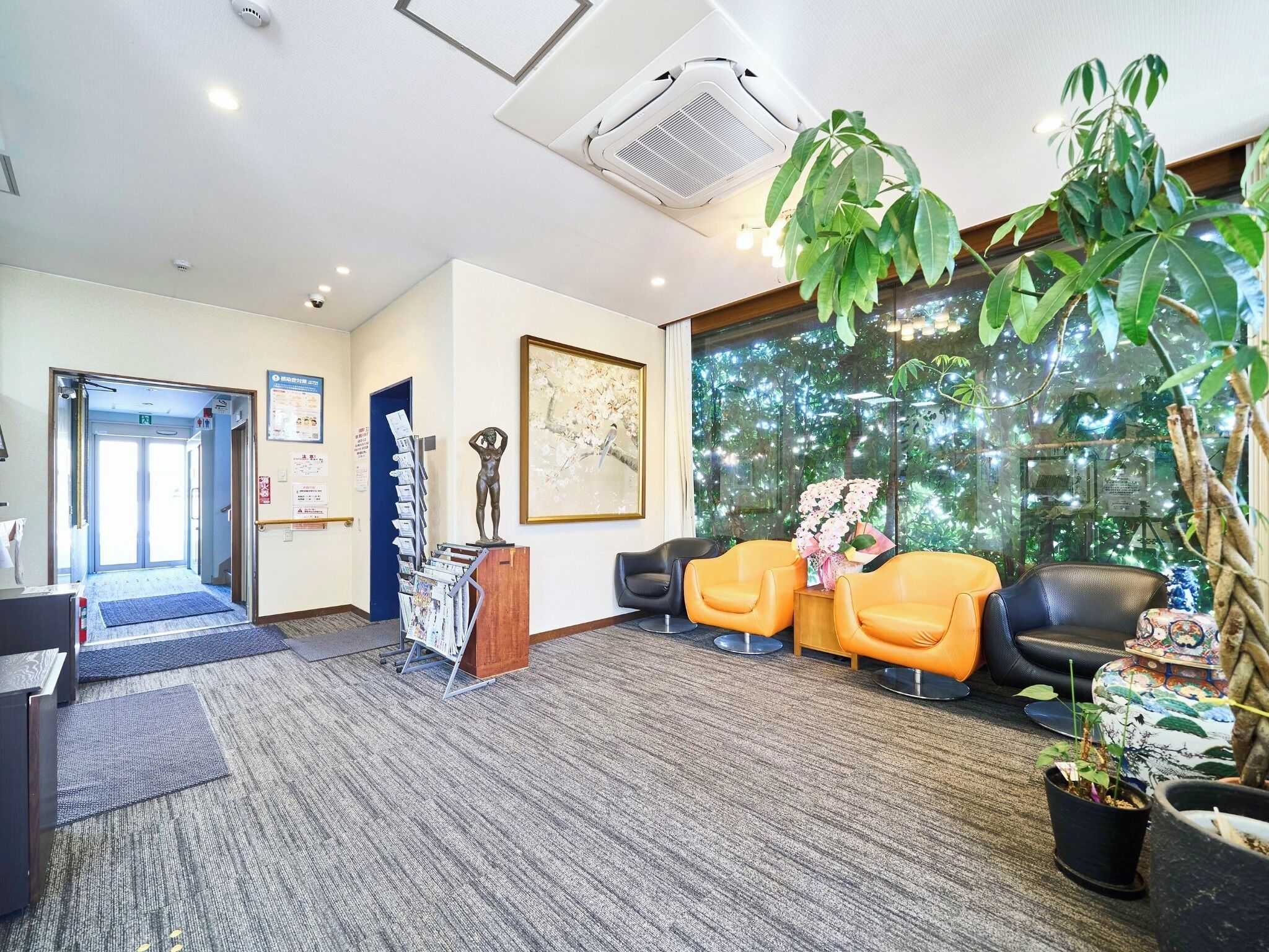 A reception area with a desk, two chairs, a statue, a framed picture, and a large window with a view of trees.