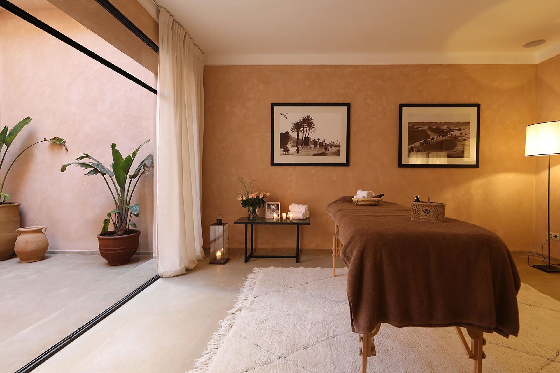 A spa room with a massage table, potted plants, framed pictures, and a lamp.