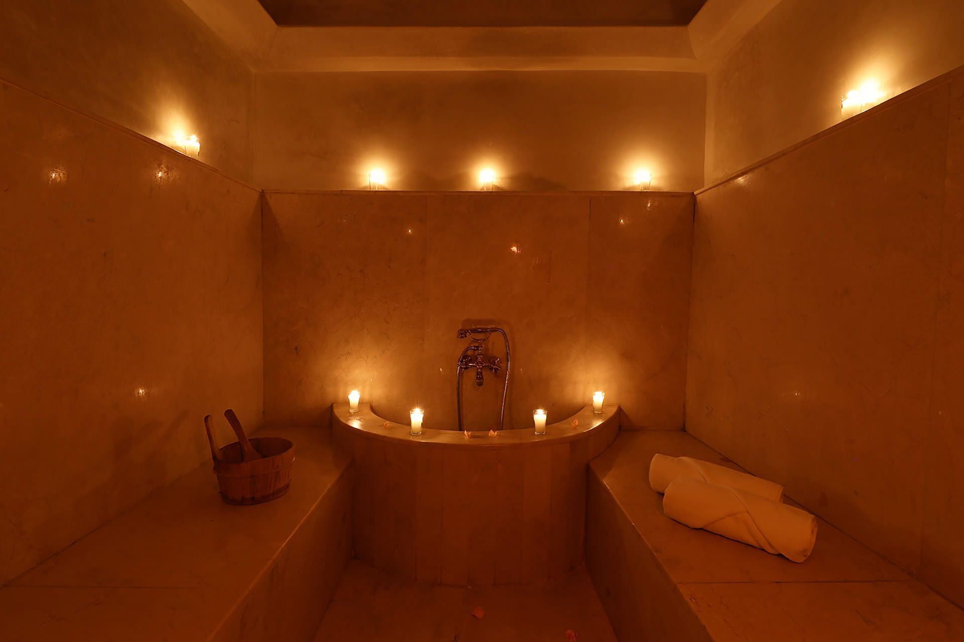 A dimly lit, circular bathtub with candles, a wooden bucket, and a towel on a bench.