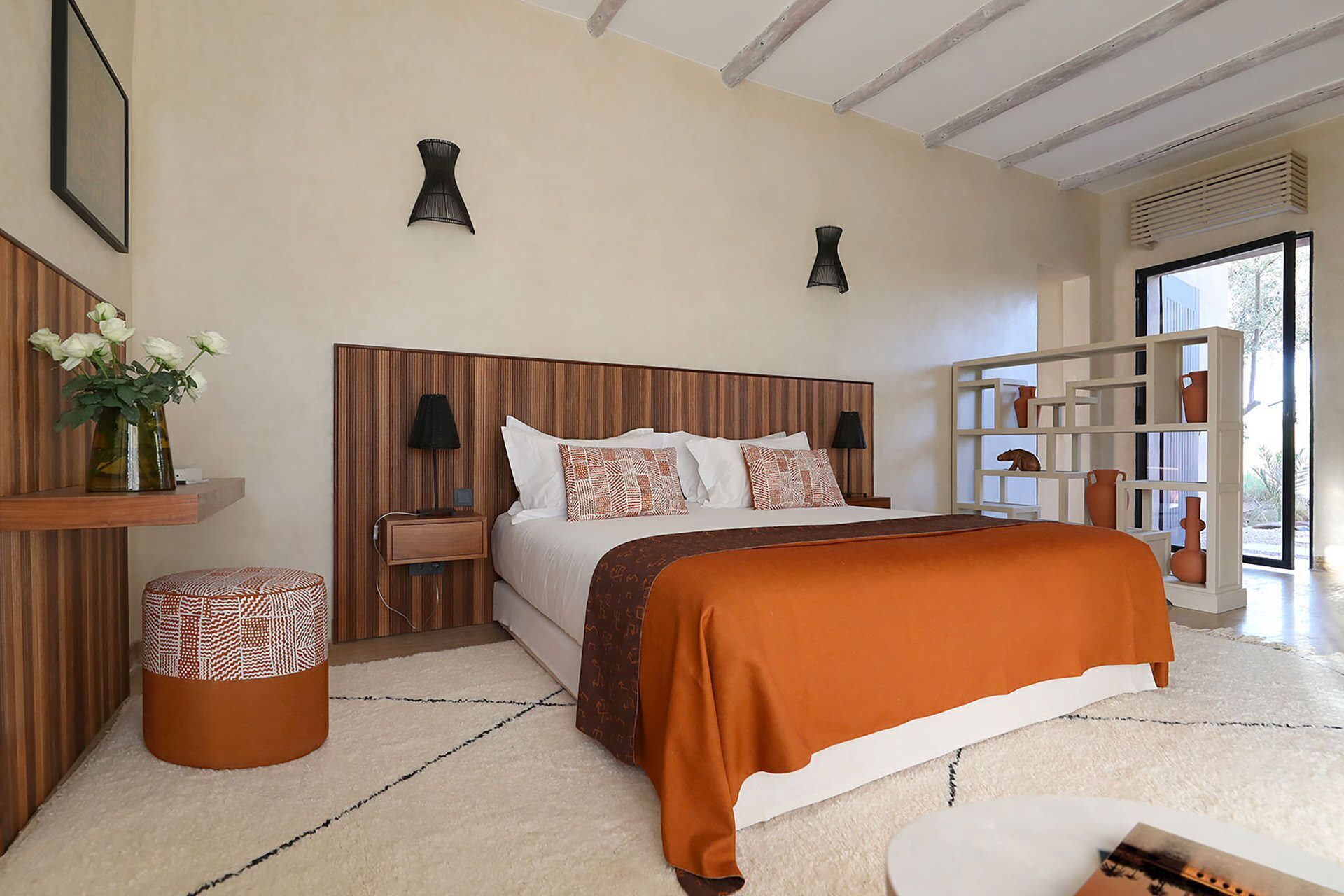 Une chambre à coucher avec une tête de lit en bois, un grand lit recouvert d’une courtepointe orange, une table de chevet avec une lampe, un petit pouf rond et une vue sur l’extérieur grâce à une porte vitrée.