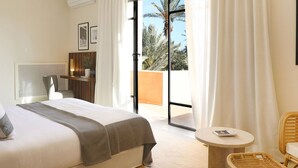Ein Schlafzimmer mit Bett, Schreibtisch, Stuhl und Blick auf Palmen durch das Fenster.