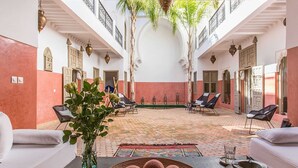 A spacious courtyard with a tiled floor, surrounded by white walls and red accents, featuring lounge chairs and a central seating area. - My Rental Homes | Riad Special with two courtyards and  swimmingpool (Marrakesh)