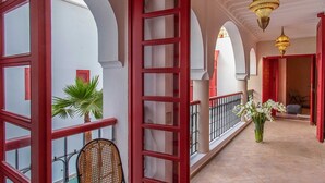 Um corredor com janelas emolduradas em vermelho, uma cadeira de vime, uma planta em vaso e um lustre.