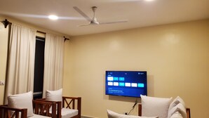 Ein Hotelzimmer mit Deckenventilator, einem wandmontierten Flachbildfernseher und zwei Holzstühlen mit weißen Kissen.